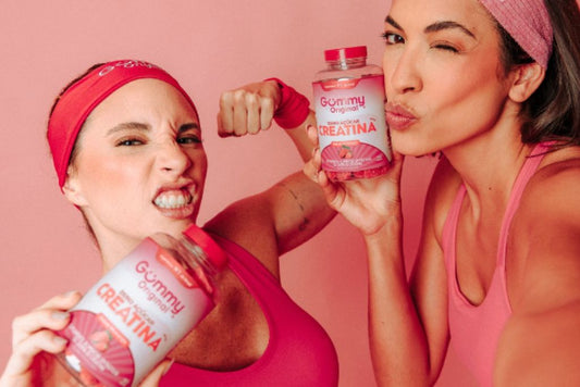 Duas mulheres com roupas esportivas segurando potes de creatina em goma da Gummy® Original. Uma delas faz pose de força e a outra pisca para a câmera. Fundo rosa vibrante.