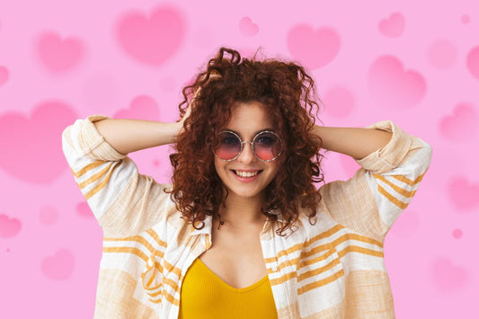 Mulher sorridente com cabelo cacheado solto, usando óculos de sol, em fundo rosa com corações representandocronograma capilar para cabelocacheado