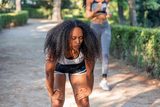 Mulher negra com cabelo cacheado aparenta cansaço após corrida ao ar livre, curvada com as mãos nos joelhos. A cena ilustra sintomas comuns da vitamina D baixa, como fadiga persistente e fraqueza muscular.