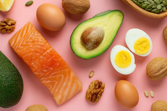 Vista superior de alimentos ricos em vitamina E, como salmão, abacate, ovos e castanhas, dispostos sobre fundo rosa, ilustrando as fontes naturais em que os benefícios da vitamina E podem ser aproveitados. 